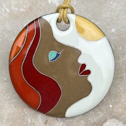 Pendentif rond en émaux sur cuivre cloisonnés qui représente le visage d'une femme de profil. Ses cheveux sont roux, ses yeux sont fermés et sa paupière est maquillé de bleu, ses lèvres sont rouge. Sa tête est penchée sur l'arrière face au soleil.