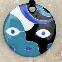 Pendentif rond en émaux sur cuivre cloisonnés. Il représente la tête d'un personnage. Les émaux sont bleu, noir et blanc, inspirés du papillon morpho.