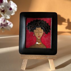 Petit tableau en émaux sur cuivre cloisonnés représentant le visage d'une femme qui ferme les yeux. Sa chevelure est un carré court frisé et ses cheveux sont noirs. Elle porte aussi des perles en boucles d'oreilles et un collier de trois perles : une blanche, une jaune et une orange. Le cadre est en cuir noir.