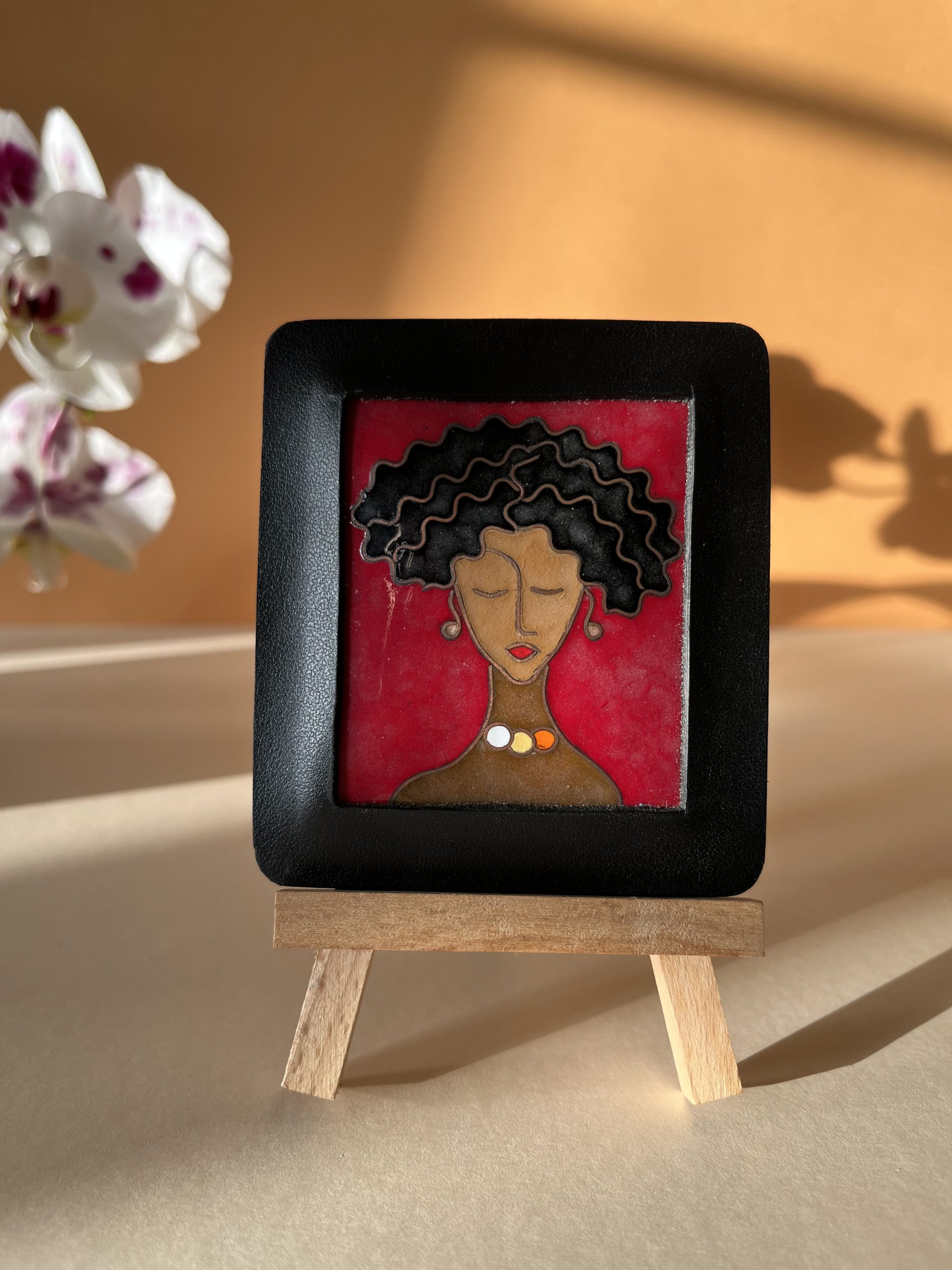 Petit tableau en émaux sur cuivre cloisonnés représentant le visage d'une femme qui ferme les yeux. Sa chevelure est un carré court frisé et ses cheveux sont noirs. Elle porte aussi des perles en boucles d'oreilles et un collier de trois perles : une blanche, une jaune et une orange. Le cadre est en cuir noir.