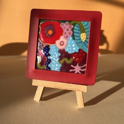 Petit tableau en émaux sur cuivre présenté sur un chevalet en bois posé sur une table. Le tableau représente des fleurs sauvages multicolores. Il est encadré dans un cuir rouge.