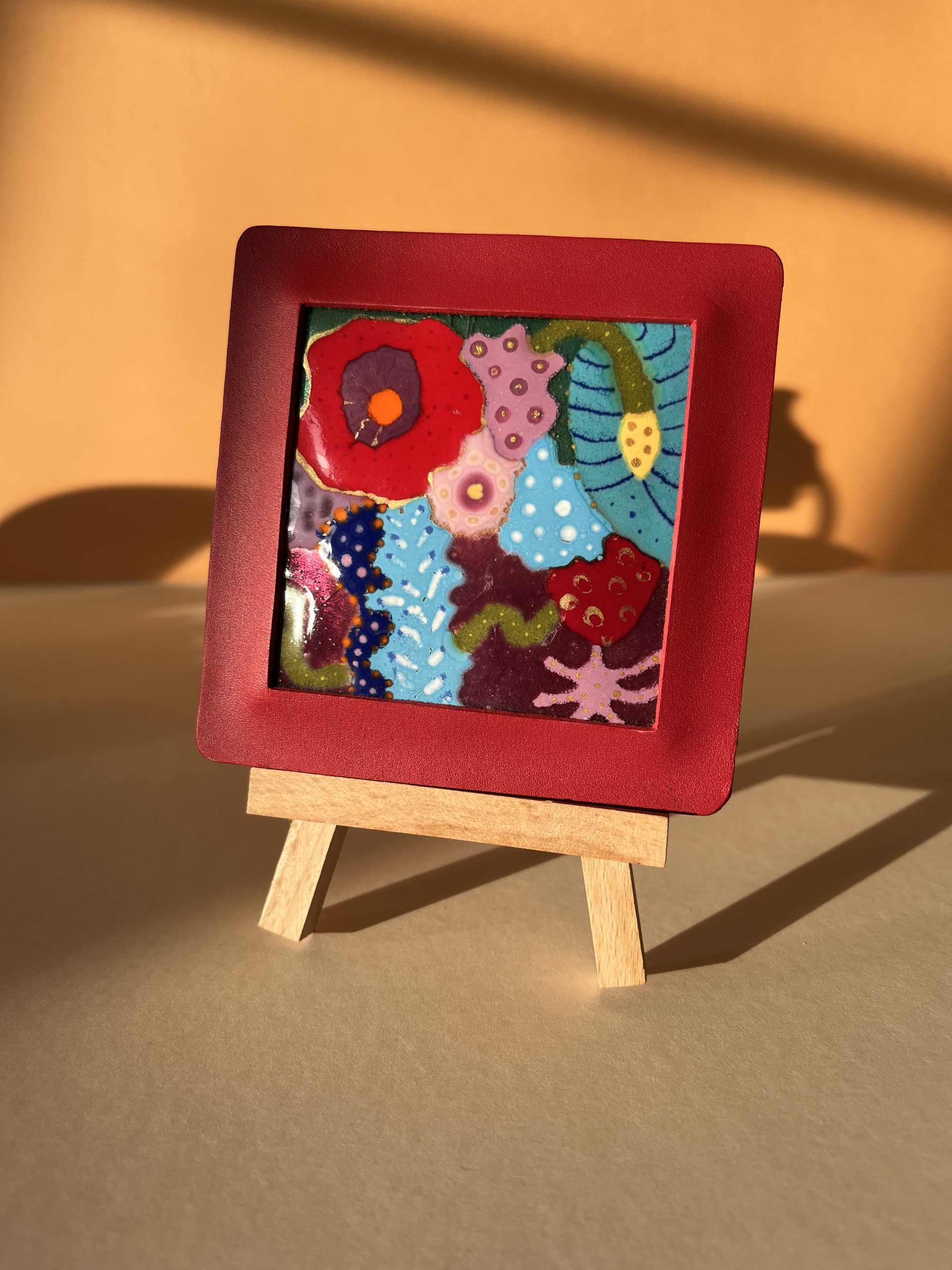Petit tableau en émaux sur cuivre présenté sur un chevalet en bois posé sur une table. Le tableau représente des fleurs sauvages multicolores. Il est encadré dans un cuir rouge.